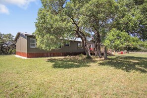A single-story house with a large tree in front, a fenced yard, and a clear sky.