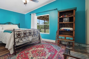 A bedroom with a bed, a window, a bookshelf, and a patterned rug. - Tejas Hills | 4BR Retreat with Firepit & Hill View (Pipe Creek)