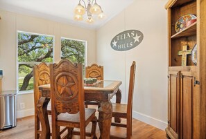 A rustic dining area featuring a heavy, hand-carved wooden table and chairs adorned with large Texas stars, set against a backdrop of a "Tejas Hills" wall sign and views of lush greenery through the windows. - Tejas Hills | 4BR Retreat with Firepit & Hill View (Pipe Creek)