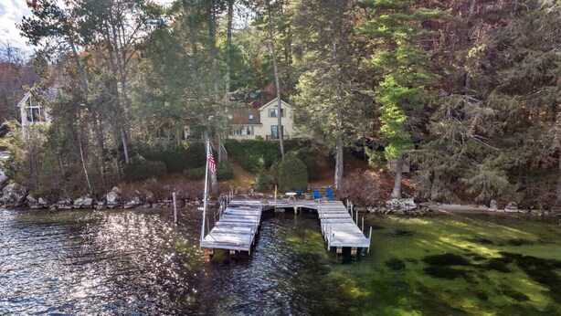 A dock extending into a calm lake surrounded by dense trees, with a house visible in the background. - Chestnut Cove Cottage- Winnipesaukee - Alton (Alton)