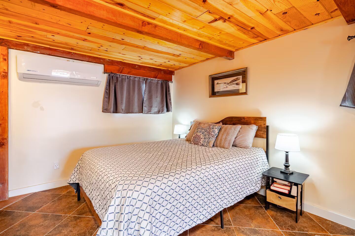 A bedroom with a wooden ceiling, a bed with patterned bedding, a nightstand with a lamp, and a window with curtains.