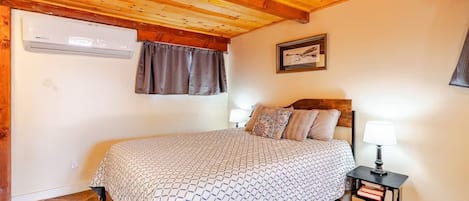 A bedroom with a wooden ceiling, a bed with patterned bedding, a nightstand with a lamp, and a window with curtains.