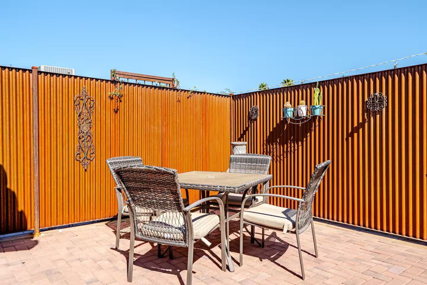 Un patio al aire libre con una mesa y cuatro sillas de mimbre, una valla metálica decorativa y una unidad de aire acondicionado pequeña fijada en la valla.