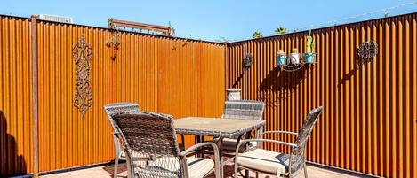 Un patio al aire libre con una mesa y cuatro sillas de mimbre, una valla metálica decorativa y una unidad de aire acondicionado pequeña fijada en la valla.