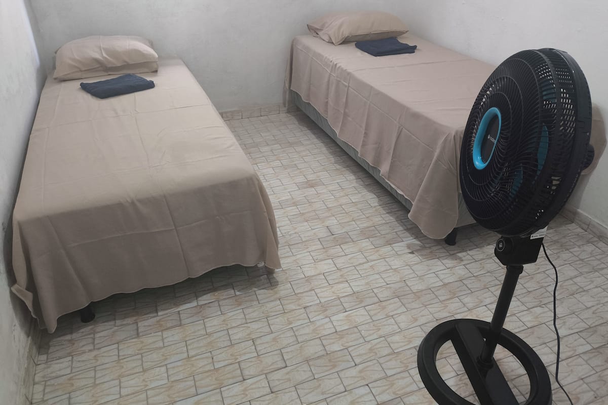 A small, clean room with two single beds, a standing fan, and a window.