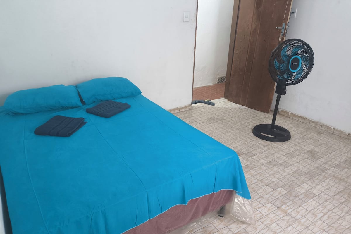 A room with a bed covered in a blue bedspread, a black fan, and a door.