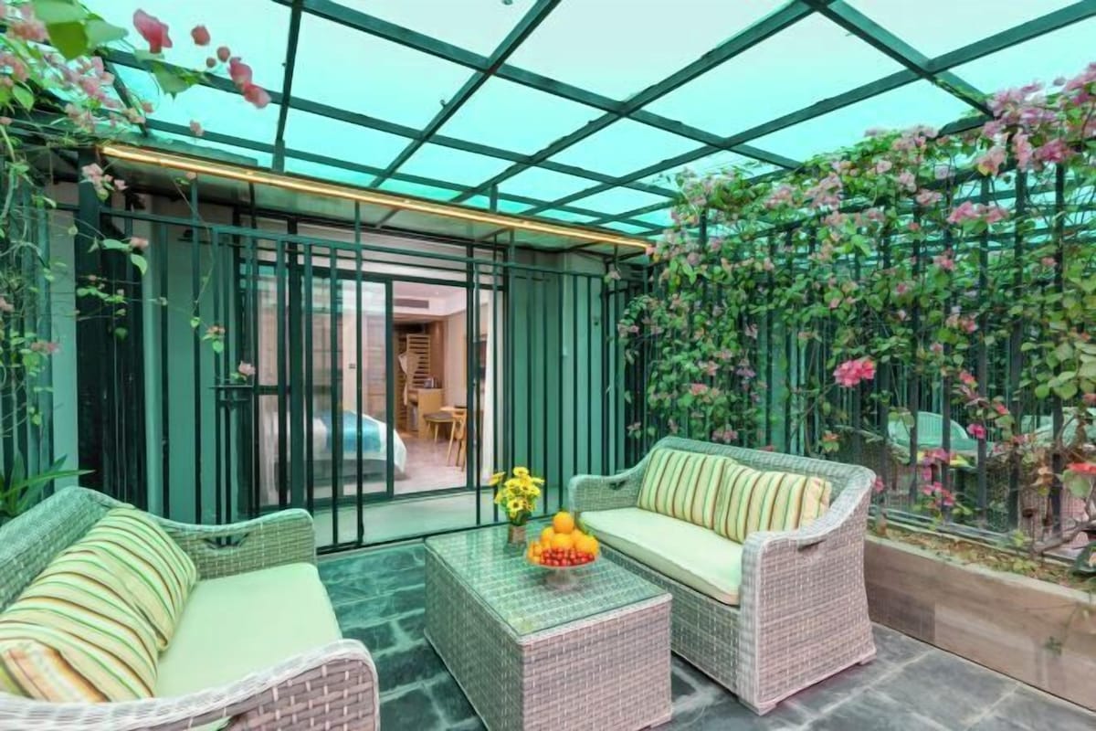 Une véranda meublée de meubles en osier, avec une table basse en verre et une vue sur un jardin où poussent des plantes grimpantes.