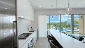 A modern kitchen with a large island, stainless steel appliances, and a view of a pool and palm trees. - Modern Luxury 2bed by 7 Mile Beach (George Town)