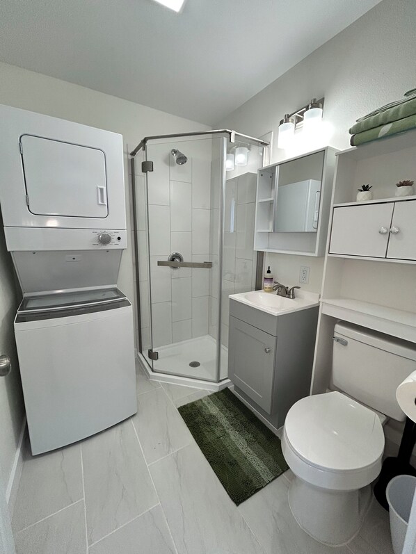 A modern bathroom with a built-in washer and dryer, a glass shower enclosure, a sink with a mirror, and a toilet. - Cozy Cody Getaway (Cody)