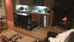A cozy living room with a fireplace, a dining area, and a kitchen with wooden cabinets and a microwave. - Golf course condo, 10 min to Torch Lake, w/pool, hot tub and sauna! (Mancelona)