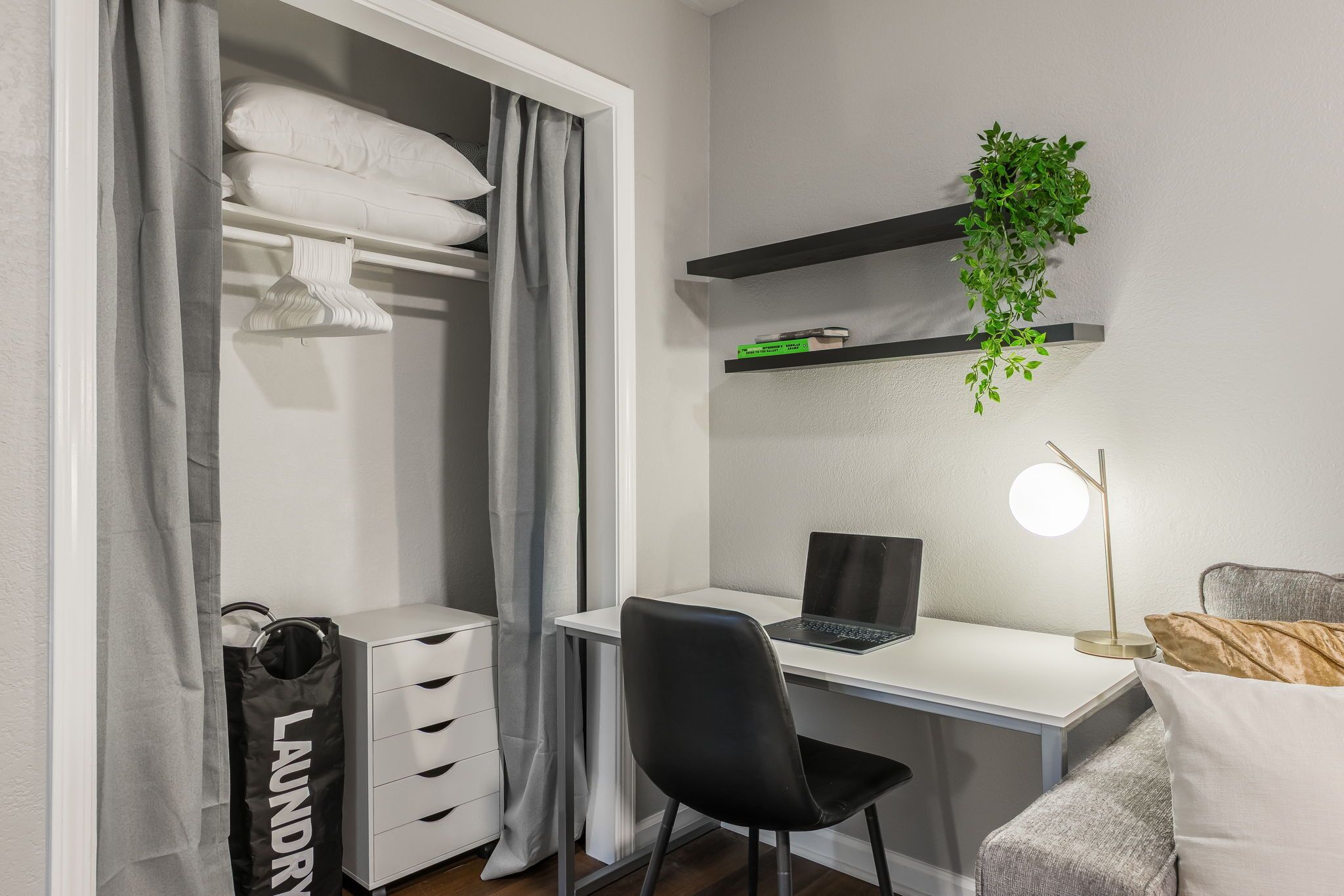 Ein ordentlich eingerichtetes Schlafzimmer mit einem weißen Schreibtisch, einem Laptop, einem schwarzen Stuhl, einer Nachttischlampe, einem grauen Sofa, einem Kleiderschrank mit Kleidung und einer Zimmerpflanze auf einem Regal.