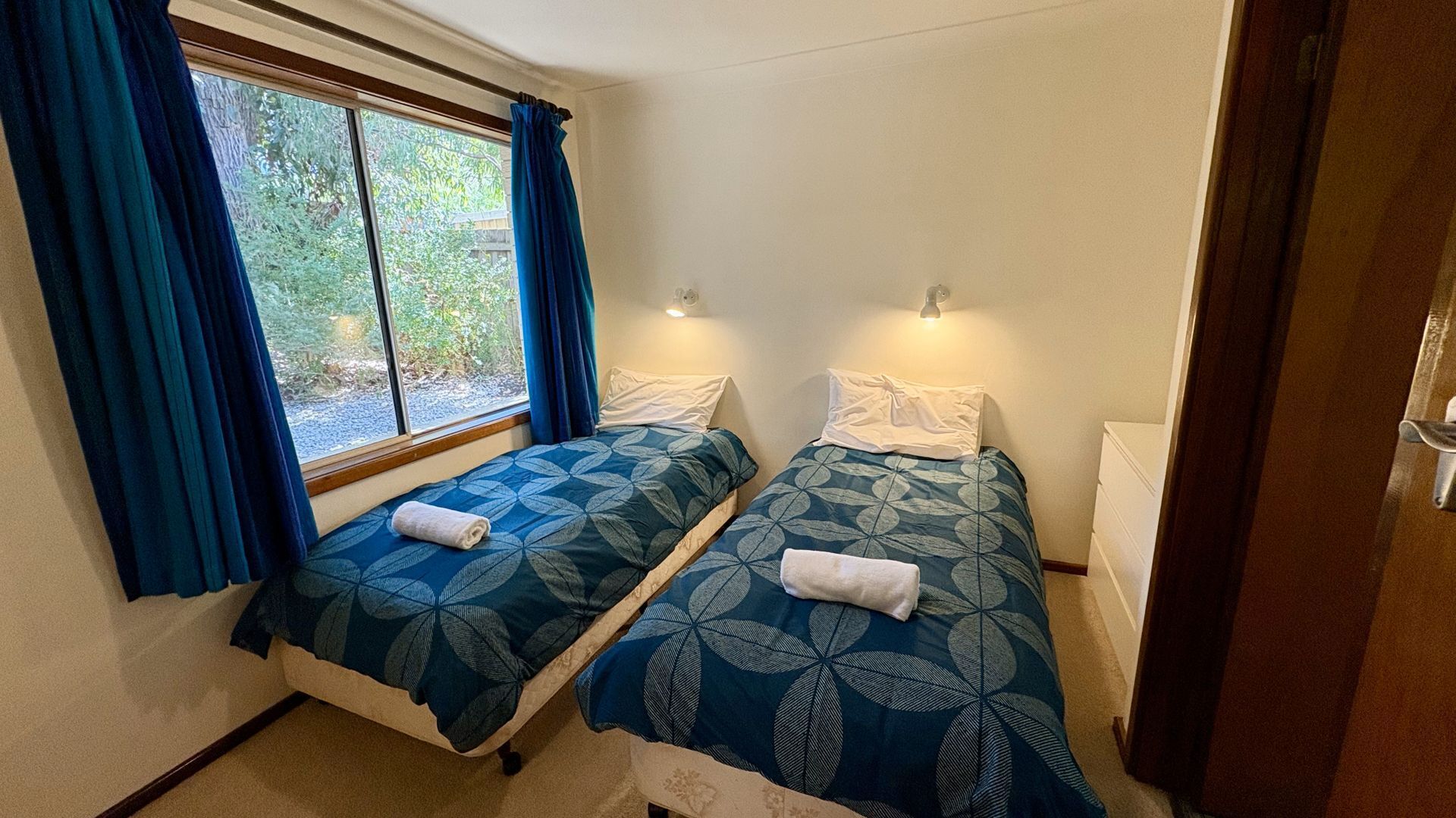 A small, cozy room with two single beds, each with a patterned duvet and pillows. There is a window with blue curtains, a bedside table with a lamp, and a door to the right.