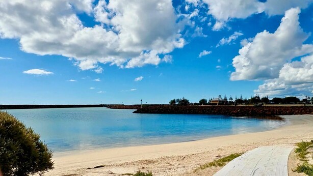 A beach with clear water, a sandy shore, and a rocky outcrop with buildings in the background.