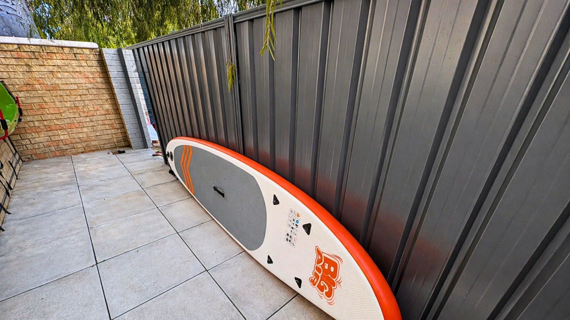 Un patio con baldosas y una tabla de surf apoyada contra una valla metálica.
