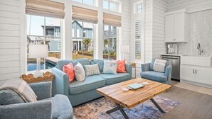 Ein geräumiges Wohnzimmer mit blauen Sofas, einem Holztisch und Blick auf einen Pool sowie benachbarte Häuser.