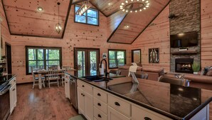 A spacious kitchen with a large island, a dining area with chairs, a fireplace, and a stone accent wall. - Open concept, 4 bd/4bath cabin in the woods, sleeps 12. Great location! (Broken Bow)