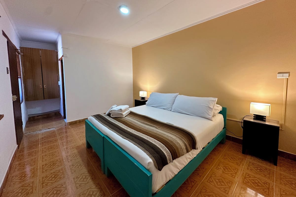 Una habitación de hotel con una cama, dos mesitas de noche con lámparas, un armario de madera y un suelo estampado.