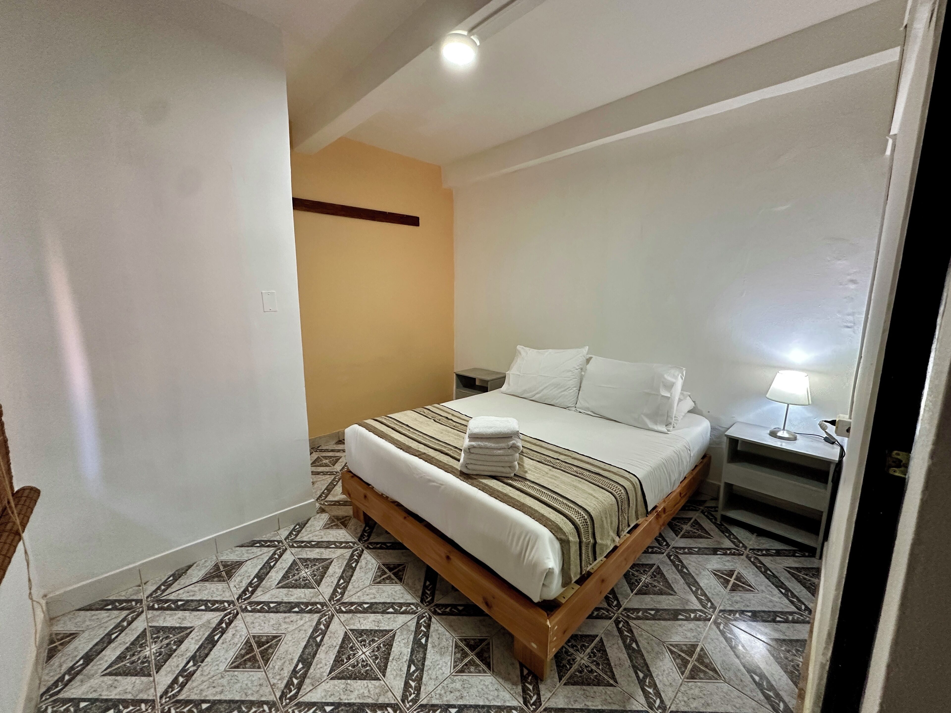 Un dormitorio con una cama, una mesita de noche, una lámpara y baldosas de suelo con diseño.