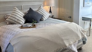 Una cama bien hecha con almohadones a rayas, una lámpara de noche y un cuadro enmarcado encima.