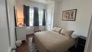 A bedroom with a bed, a chair, a desk with a lamp, a window with curtains, and a painting on the wall.