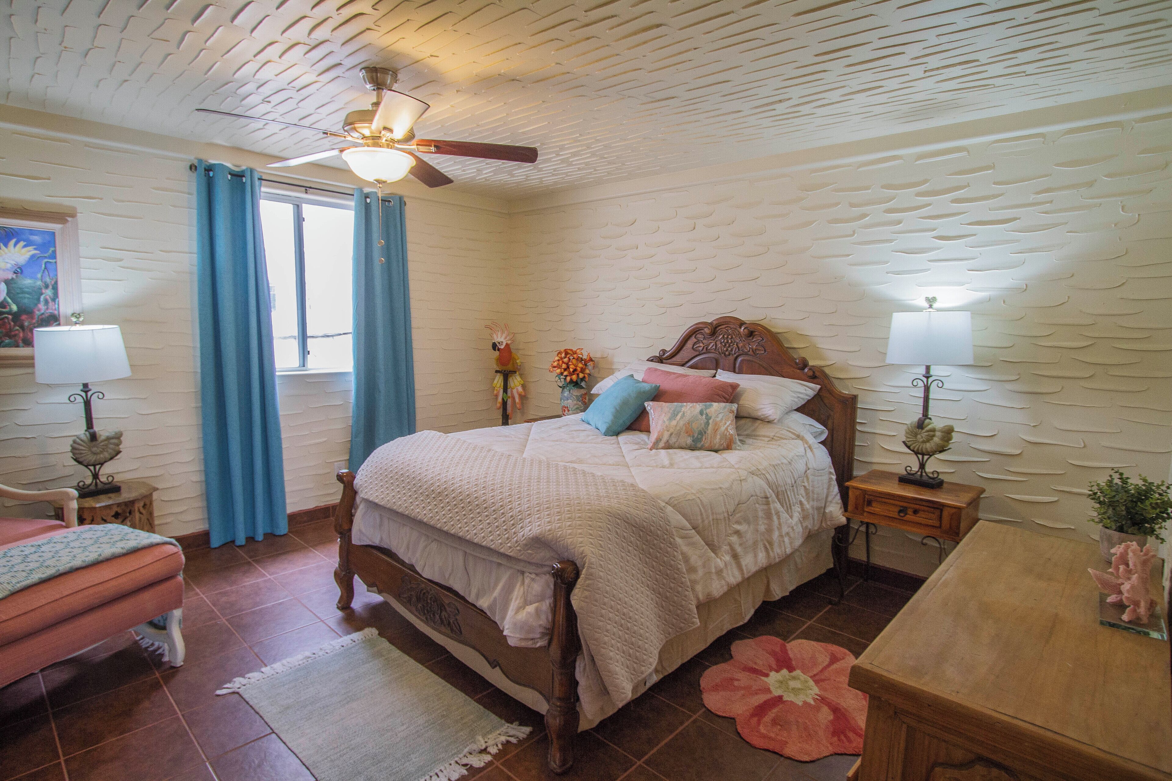 A bedroom with a wooden bed, a ceiling fan, a window with blue curtains, and a nightstand with a lamp.