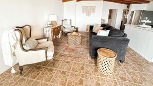 Een woonkamer met een hoge plafond, houten balken, een grijze bank, een witte fauteuil en een kleine ronde tafel.
