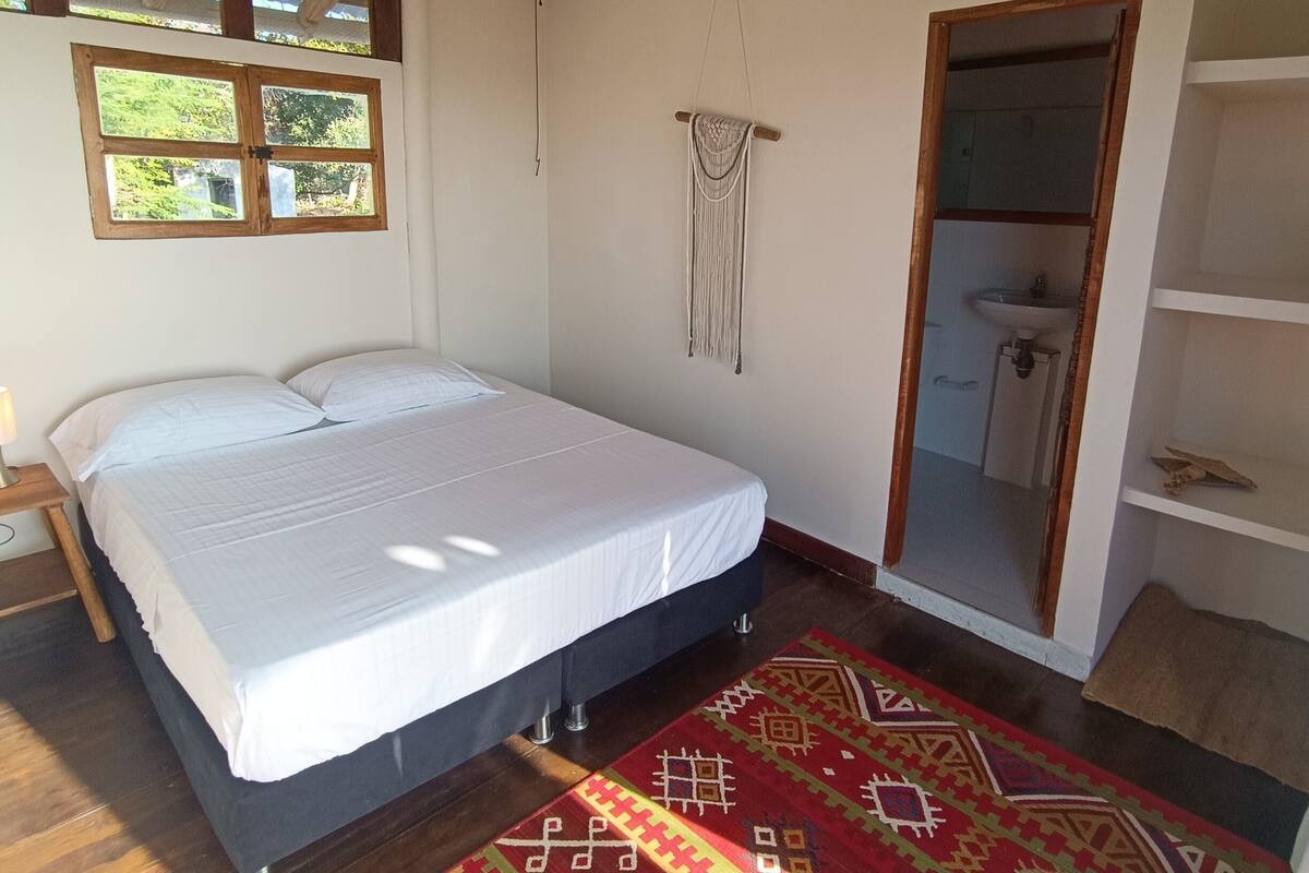 A bedroom with a bed, wooden floor, a ceiling fan, a window, and a door leading to a bathroom.