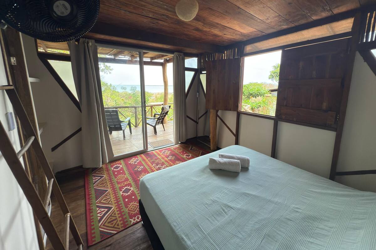 A room with a bed, a fan, a view of the outdoors, and a wooden ceiling.
