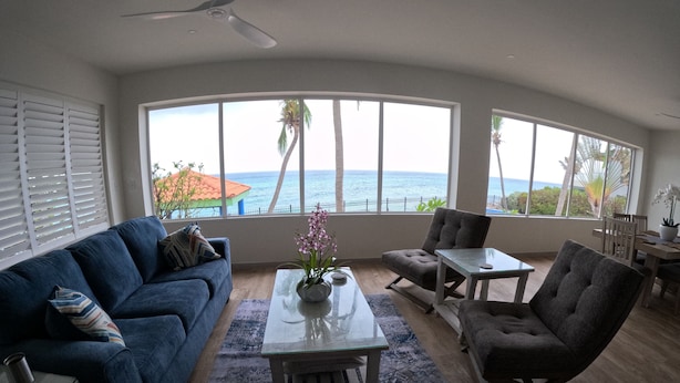 A living room with a blue sofa, a glass coffee table with a vase, a grey armchair, and a view of the ocean through large windows. - Welcome to the Dutch side of St. Maarten. 3 bedroom unforgettable ocean view. (Koolbaai)