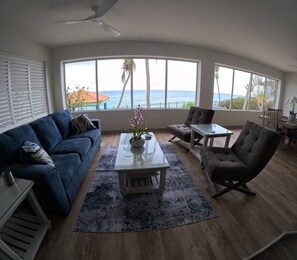 Een woonkamer met een blauwe bank, een glazen salontafel met een vaas, een grijze fauteuil en uitzicht op de oceaan door grote ramen.