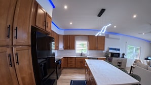 A modern kitchen with wooden cabinets, a black refrigerator, a white countertop, and a dining area with a green chair. - Welcome to the Dutch side of St. Maarten. 3 bedroom unforgettable ocean view. (Koolbaai)