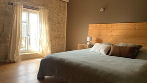 A bedroom with a stone wall, wooden ceiling beams, a bed with pillows, a bedside table with a lamp, and a window with curtains. - Exceptional group gîte for families, friends, professionals 24 pers. (Lingèvres)