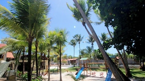 A playground with slides and swings, surrounded by palm trees and a sandy ground.