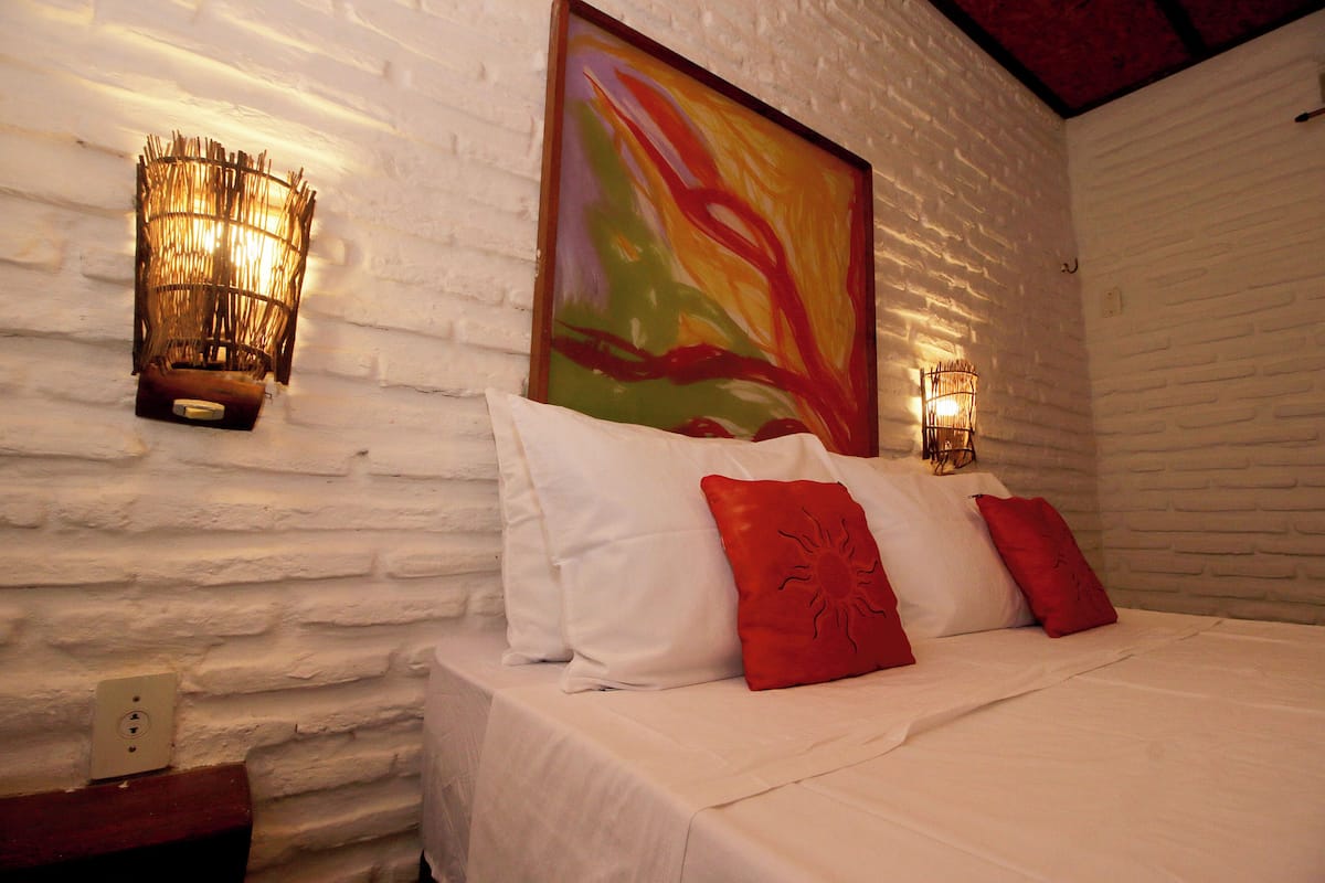 Una cama con ropa de cama blanca y almohadas rojas, un cuadro abstracto y colorido encima, y dos lámparas de pared a cada lado.