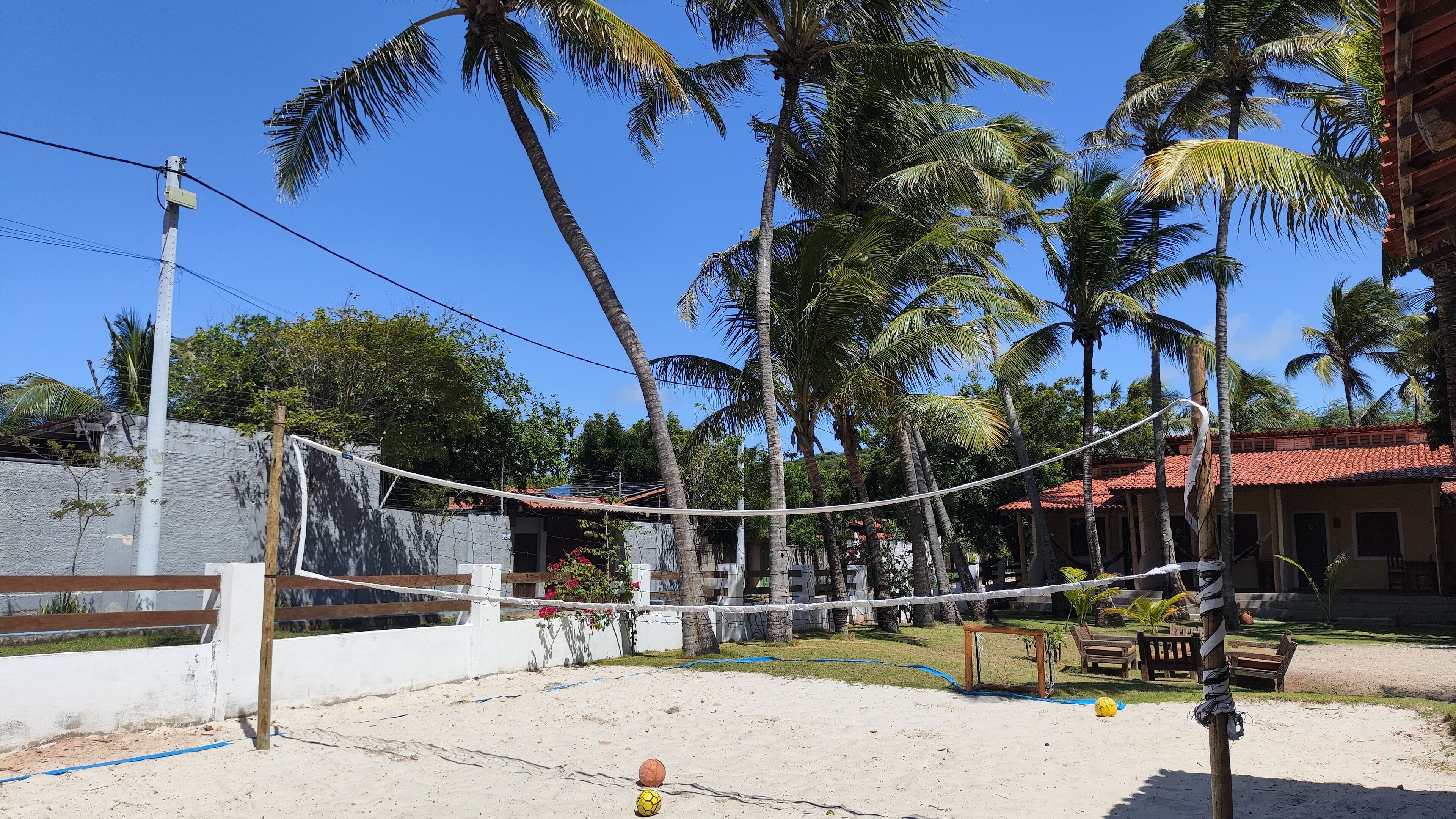 A sandy outdoor area with a volleyball net, palm trees, and a building with a red roof.