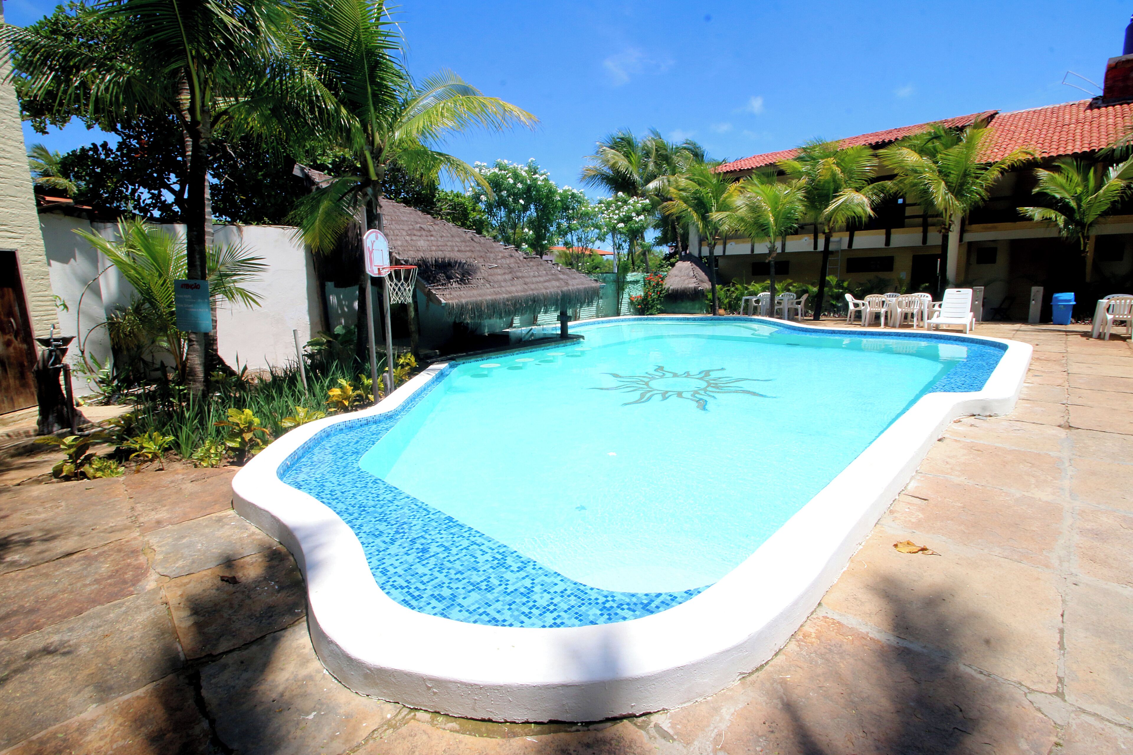 A rectangular swimming pool with a blue mosaic border, surrounded by palm trees and a building with a red-tiled roof.