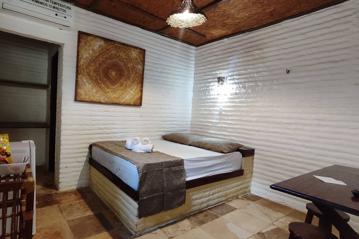Una habitación pequeña y acogedora con una cama, una mesa y una silla. Hay un cuadro en la pared y un cartel sobre la cama.