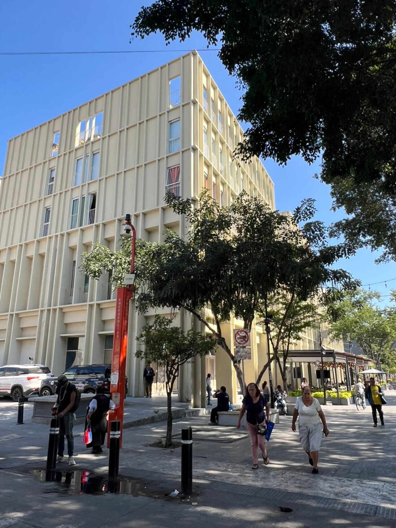 Um edifício de vários andares com fachada em estilo grade, caixas de emergência e uma área para pedestres com árvores e bancos.