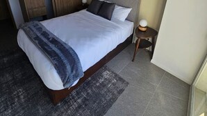 A modern bedroom with a large bed, a wooden wardrobe, a small bedside table, and a large window.