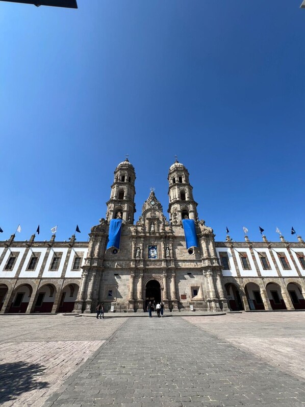 A historic building with two prominent towers, a central clock, and a paved plaza.