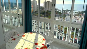 A room with a large window offering a view of the sea and buildings. There is a round table with chairs and a floral tablecloth. - Ocean Panoramic View at Playa Las Americas (Playa de la Américas)