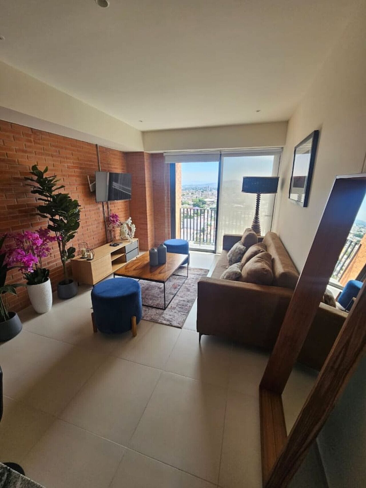 Een moderne woonkamer met een bank, salontafel en televisie. Er staan potplanten en er is een balkon met uitzicht.