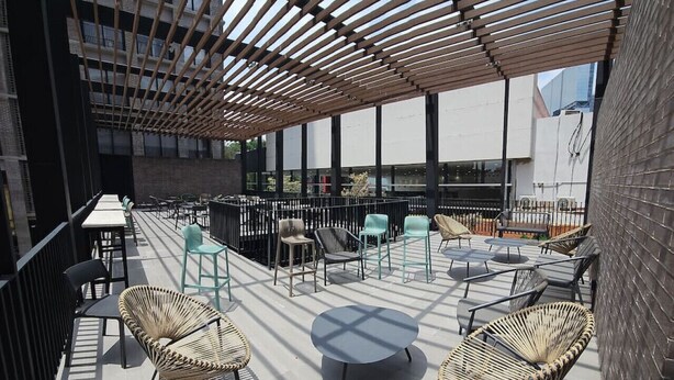 An outdoor seating area with wicker chairs and tables, a wooden pergola, and a view of a building with large windows.