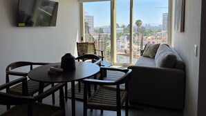 A modern living room with a sofa, a dining table with chairs, and a large window offering a city view.