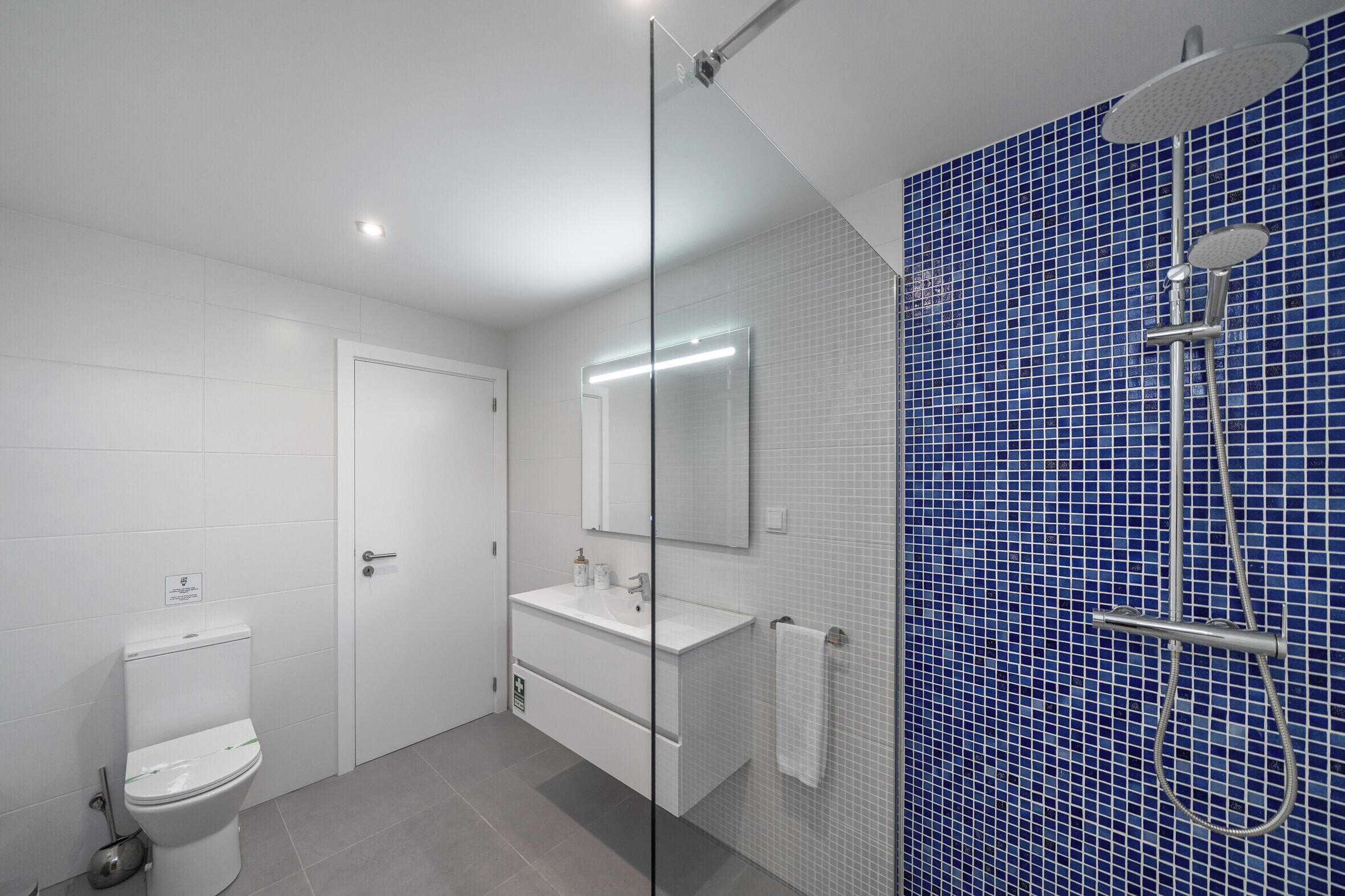 A modern bathroom with a toilet, a sink, a mirror, and a shower area with blue tiles.