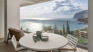 A modern dining area with a round table, chairs, and a view of the sea and coastline. - With pool and sea view - Acqua Residence III (Funchal)