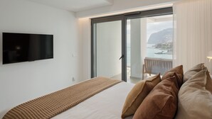 A modern hotel room with a large bed, a flat-screen TV mounted on the wall, and a balcony with a view of the sea and mountains. - With pool and sea view - Acqua Residence III (Funchal)