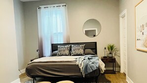 A bedroom with a bed, two ottomans, a bedside table with a plant, a mirror, and a painting on the wall. - Historic Garden 1-bedroom apartment in amazing Augusta - The Pearl with AC, WiFi (Augusta)