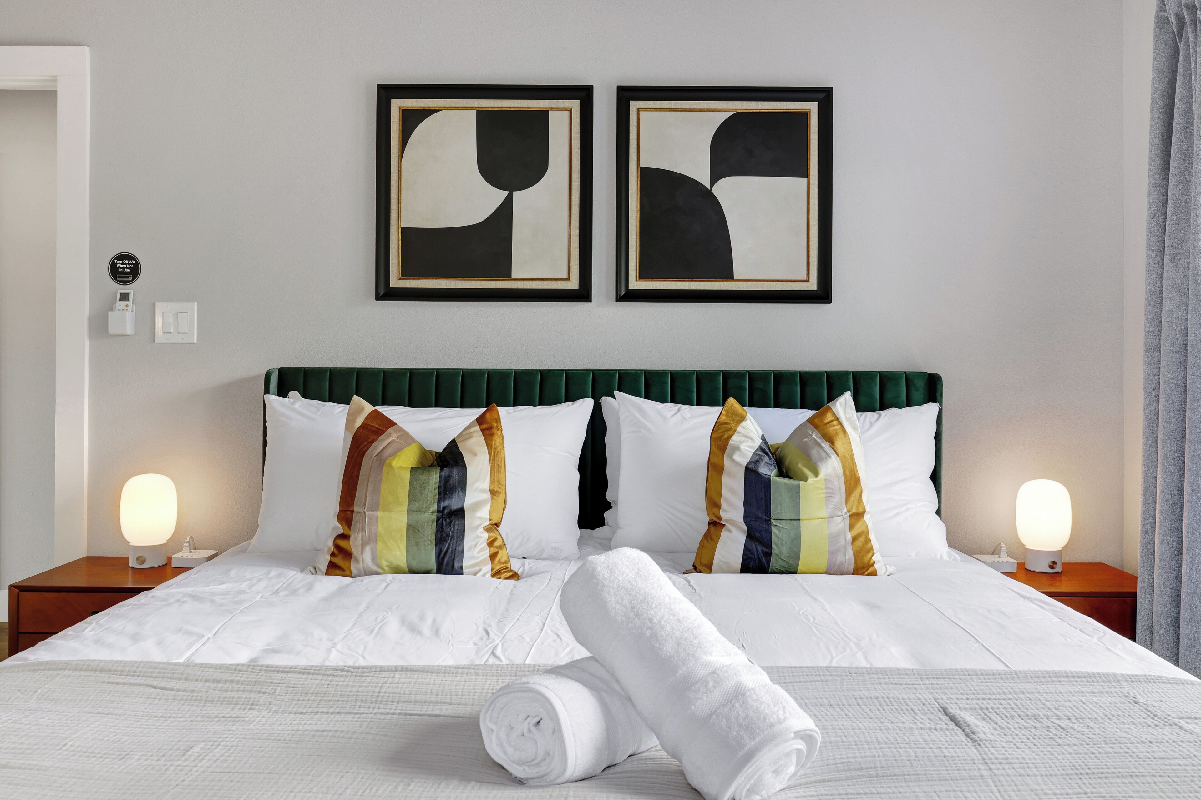 A neatly made bed with white linens, two pillows with striped patterns, and a green upholstered headboard. There are two bedside tables with lamps, and two framed abstract paintings on the wall.