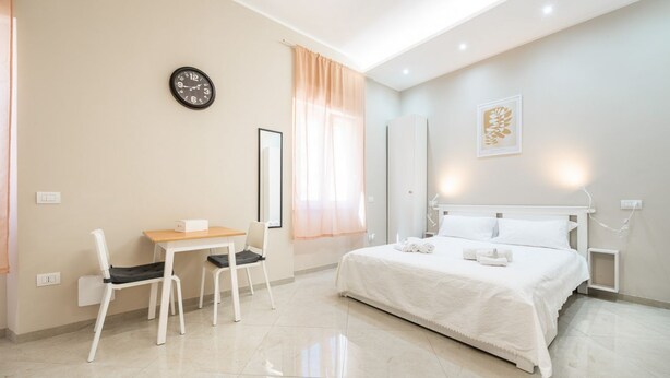 Quarto de hotel moderno com cama, escrivaninha, cadeiras, relógio e janela com cortinas.